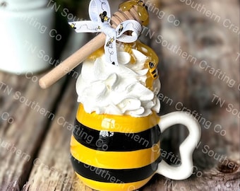 May include: A yellow and black striped ceramic mug with a white handle and a white whipped cream topping. A wooden honey dipper with a yellow honey drizzle is placed on top of the whipped cream. The mug is decorated with small bee images.