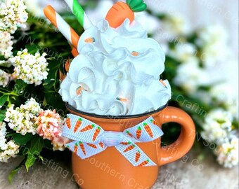 May include: Orange ceramic mug with a white whipped cream topping, a carrot on top, and a white ribbon with a carrot print. The mug is sitting on a wooden surface with white flowers in the background.