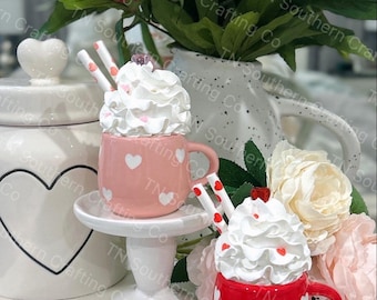 May include: Two miniature ceramic mugs with whipped cream and red and pink hearts. The mugs are on a white pedestal and a white surface with white flowers.