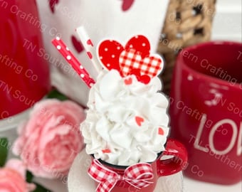 May include: A miniature red mug with a white rim and a red and white gingham bow sits on a white wooden base. The mug is filled with white whipped cream and red and white heart-shaped sprinkles. Two red and white striped straws stick out of the whipped cream.