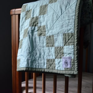 May include: A baby quilt with a patchwork design in shades of blue and gray, draped over a wooden crib. The quilt features a label that reads "FRESHWATER QUILTS". The quilt is a soft, handmade item.