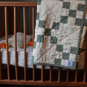 May include: A white and green patchwork baby quilt with a label that says "Freshly Picked". The quilt is draped over a wooden crib with a white and brown patterned sheet.