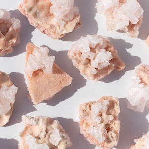 May include: A collection of pink salt crystals with white salt formations on top. The salt crystals are arranged on a white surface.