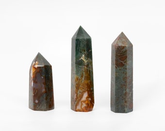 May include: Three green, brown, and orange agate crystal points. The points are different sizes and are arranged in a row on a white surface.