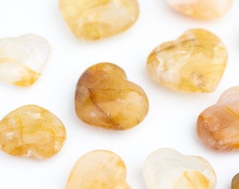 May include: A collection of heart-shaped, clear, yellow-tinted quartz crystals with natural inclusions. The crystals are arranged on a white surface.