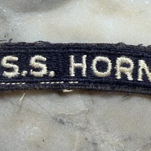 May include: A rectangular embroidered patch with a dark navy blue background and white stitched lettering that reads U.S.S. HORNET. The patch features a finished border with white thread detailing and shows signs of wear along the edges.