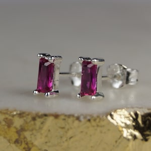 May include: A pair of silver stud earrings featuring rectangular, magenta-coloured gemstones. The earrings have a simple, elegant design, highlighting the bright colour of the stones. The studs are displayed on a light-coloured surface.