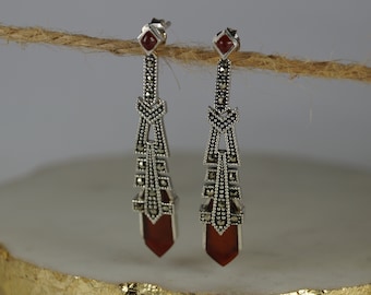May include: A pair of silver drop earrings with a geometric design. Each earring has a red gemstone at the top and bottom, with marcasite accents. The earrings are suspended from a silver post.