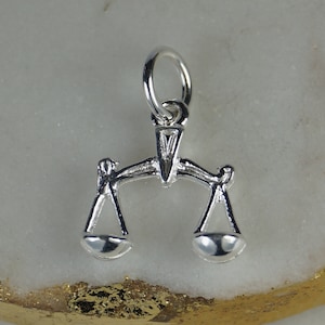 Peut inclure: Un petit charm argenté brillant en forme de balance, souvent associé à la justice. Le charm est doté d'une boucle pour l'attacher à un collier ou un bracelet. Les plateaux de la balance sont équilibrés.