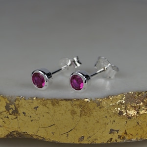 Puede incluir: Un par de pendientes de plata con gemas rosas brillantes y redondas. Las gemas están engastadas en un bisel. Los pendientes se muestran sobre una superficie dorada y blanca. Un artículo de joyería clásico.
