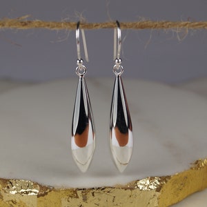 May include: A pair of polished silver teardrop earrings suspended from a natural fibre string. The earrings have a smooth, reflective surface and a simple, elegant design. The background is a neutral colour.
