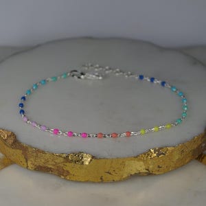 Peut inclure: Un bracelet en argent orné de petites perles colorées. Les perles présentent un dégradé arc-en-ciel, allant du bleu au violet, rose, orange et jaune. Le bracelet est présenté sur une surface dorée et blanche.