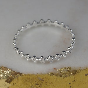 Op de afbeelding: Een zilveren ring met een kralenontwerp. De ring heeft een reeks kleine, ronde kralen die een textuurpatroon creëren. De ring wordt getoond op een wit en goud oppervlak.