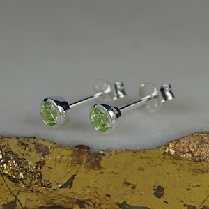 May include: A pair of stud earrings with round, light green gemstones set in silver bezels. The earrings are displayed on a gold and white surface, showcasing their bright, polished appearance. The earrings are a classic jewelry item.