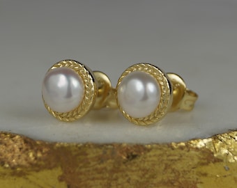 May include: A pair of gold stud earrings with a round, white pearl center. The pearl is surrounded by a textured, gold border. The earrings are displayed on a gold and white surface.