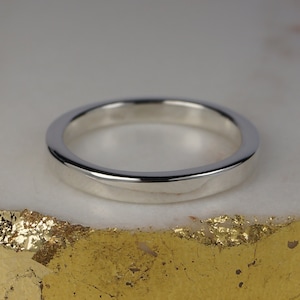 May include: A polished silver band ring. The ring has a flat profile and a smooth, reflective surface. The ring is set against a white and gold background. This ring is suitable for everyday wear.
