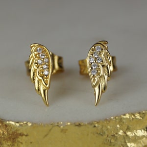 May include: A pair of gold-coloured stud earrings, each shaped like a wing. The wings are detailed with small, sparkling clear stones. The earrings are displayed on a white and gold surface.