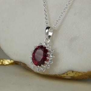 Könnte beinhalten: Eine silberne Anhängerhalskette mit einem großen, oval geschliffenen, tiefroten Edelstein, der von kleineren, klaren Edelsteinen umgeben ist. Der Anhänger hängt an einer zarten Silberkette. Der Schmuck wird auf einer hellen Oberfläche präsentiert.