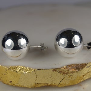 May include: A pair of spherical, silver stud earrings with a polished finish. The earrings reflect light, creating a bright appearance. They are presented on a gold and white surface. These classic earrings are a timeless jewellery accessory. The earrings are approximately 1.5 cm in diameter.