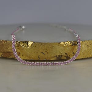 May include: A delicate pink gemstone tennis bracelet. The bracelet features a row of small, round pink gemstones set in a silver-toned metal. The bracelet is displayed on a gold-coloured surface.