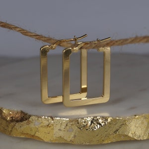 May include: A pair of gold square hoop earrings. The earrings are presented on a white and gold surface, with a natural rope in the background. The earrings have a simple, modern design.