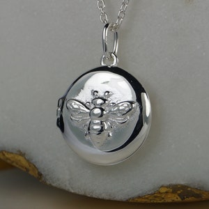 May include: A silver round locket pendant featuring a detailed bee design. The locket hangs from a delicate silver chain. The bee is embossed on the locket's surface, adding a textured element to the jewellery. The locket is a classic piece.