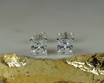 Könnte beinhalten: Ein Paar quadratisch geschliffene, klare Edelstein-Ohrstecker. Die Ohrringe sind in einem silberfarbenen Metall gefasst, und die Edelsteine sind facettiert, um das Licht einzufangen. Die Ohrringe werden auf einer gold-weißen Oberfläche präsentiert.
