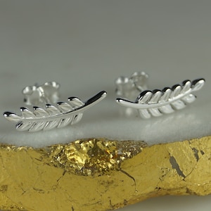 Puede incluir: Un par de pendientes de botón plateados con forma de hoja. Cada pendiente presenta un diseño detallado de hoja con múltiples segmentos. Los pendientes se muestran sobre una superficie blanca y dorada, resaltando sus cualidades reflectantes.