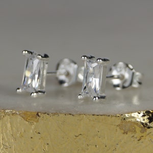 Peut inclure: Une paire de boucles d'oreilles à clous en argent avec des pierres rectangulaires en zircone cubique transparente. Les boucles d'oreilles sont serties dans un métal argenté et ont un fermoir à vis. Elles sont présentées sur une surface dorée et blanche.