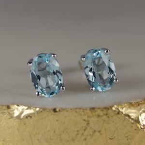 Può includere: Un paio di orecchini a lobo ovali con pietre preziose azzurre. Le pietre sono incastonate in un metallo color argento con quattro griffe. Gli orecchini sono esposti su una superficie chiara con un bordo dorato.