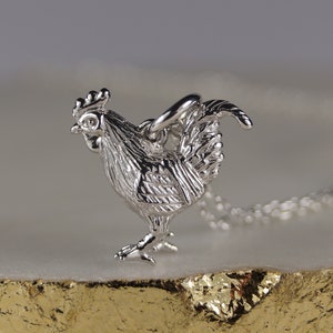May include: A silver rooster pendant on a delicate chain. The detailed charm depicts a rooster with textured feathers, a comb, and a wattle. The pendant is displayed on a white and gold surface.