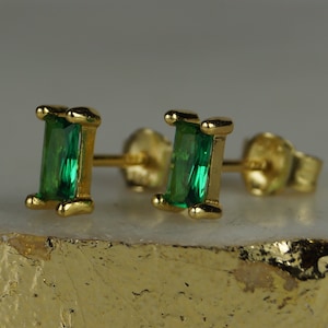 Könnte beinhalten: Ein Paar goldfarbene Ohrstecker mit rechteckigen, smaragdgrünen Edelsteinen. Die Ohrringe sind in einer goldenen Lünettenfassung gefasst und haben einen Stecker und einen Butterfly-Verschluss. Die Ohrringe werden auf einer hellen Oberfläche präsentiert.