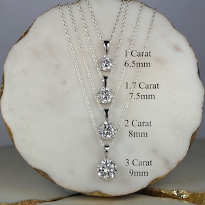 Op de afbeelding: Vier zilveren kettingen met diamantachtige hangers van verschillende maten. De hangers zijn gelabeld met hun karaatgewicht en grootte in millimeters: 1 karaat (6,5 mm), 1,7 karaat (7,5 mm), 2 karaat (8 mm) en 3 karaat (9 mm).