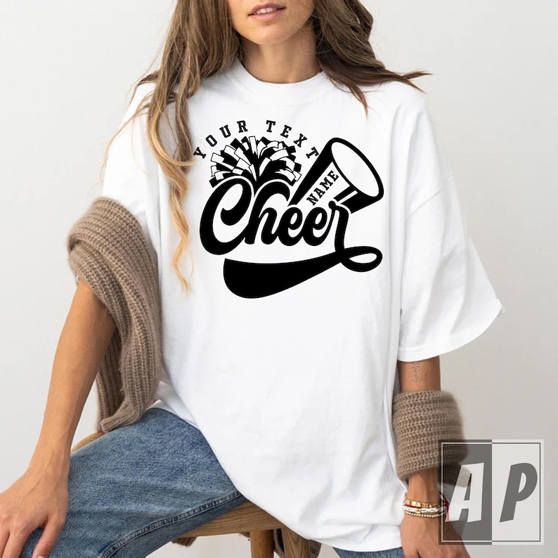 Peut inclure: T-shirt blanc surdimensionn&eacute; avec un graphique noir d'un pompon de pom-pom girl et d'un m&eacute;gaphone avec le texte "Your Text Name Cheer".
