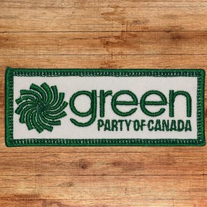 Op de afbeelding: Geborduurde patch van de Green Party of Canada met een witte achtergrond en groene letters. De patch heeft een groen gestileerd swirl-ontwerp.