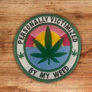 Peut inclure: Patch brodé vert avec une feuille de marijuana et le texte "Personnellement victime de mon herbe" dans un cercle blanc avec un design de coucher de soleil rose, jaune et bleu.
