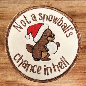 以下が含まれることがあります： 茶色の縁取りのある白い刺繍パッチ。パッチには、赤いサンタ帽をかぶり、雪玉を持っている茶色のクマの漫画が描かれています。パッチのテキストは「Not a snowball's chance in hell.」です。