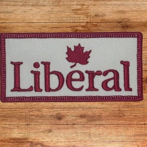 Puede incluir: Un parche blanco con una hoja de arce roja bordada y la palabra "Liberal" en rojo.