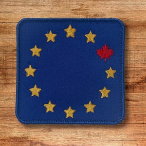 May include: A square embroidered patch featuring a dark blue background with a circular arrangement of twelve icons. Eleven of these icons are yellow five-pointed stars, while one icon is a red maple leaf positioned in the upper right quadrant.
