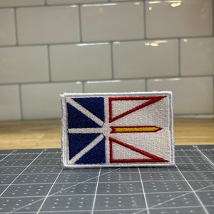 May include: A white, red, and blue embroidered patch with a yellow arrow in the center. The patch is a representation of the flag of Newfoundland and Labrador.