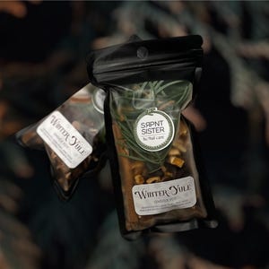 Puede incluir: Dos bolsas negras llenas de hierbas y especias, etiquetadas como "Winter Yule". Una bolsa tiene una pegatina "SARNT SISTER". El contenido incluye palitos de canela, rodajas de naranja y otros productos botánicos. El fondo es una vista borrosa de follaje verde.