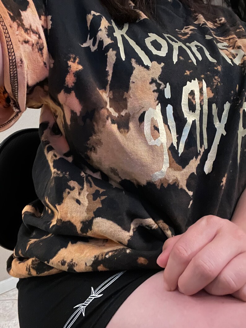 Pu&ograve; includere: T-shirt nero e beige tie-dye con la scritta argentata "Korn's Girly". Abbinato a pantaloncini neri con un disegno di filo spinato bianco.