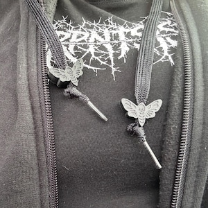 May include: A black hooded sweatshirt featuring a white graphic print on the chest. The two black drawstrings are adorned with matte black moth-shaped aglets, each featuring a detailed skull pattern on the body of the moth, positioned above the tips.