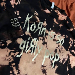 Pu&ograve; includere: T-shirt nero con effetto tie-dye e testo argentato "Korn is so girly pop". La maglietta presenta anche il testo "Srs Sis" in argento. Un capo di moda unico e audace.