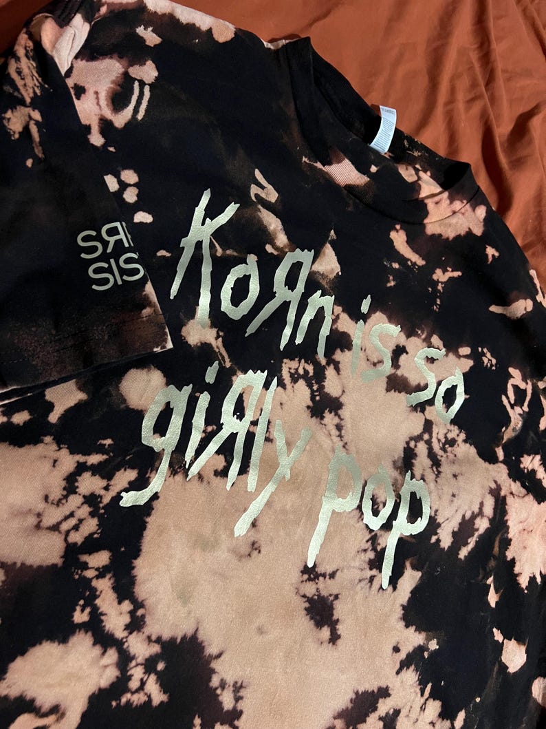 Pu&ograve; includere: T-shirt nero con effetto tie-dye e testo argentato "Korn is so girly pop". La maglietta presenta anche il testo "Srs Sis" in argento. Un capo di moda unico e audace.