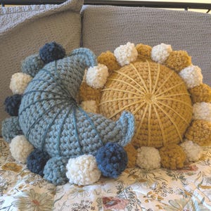 Puede incluir: Dos cojines decorativos de ganchillo. Uno tiene forma de media luna en azul con pompones azules y blancos. El otro es redondo, color mostaza, con pompones blancos y mostaza. Los cojines están sobre una colcha con estampado floral.
