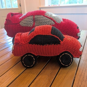 以下が含まれることがあります： 手作りのかぎ針編みの車の玩具2つ。小さい車は赤色で、黒い窓と黒い車輪が付いています。大きい車は赤色で、グレーと白のアクセントが付いています。どちらも木製の表面に置かれています。