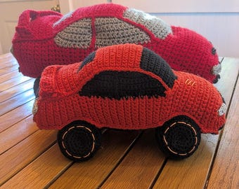 May include: Two handmade crochet car toys. The smaller car is red with black windows and black wheels. The larger car is red with gray and white accents. Both are sitting on a wooden surface.