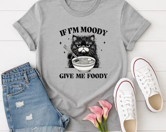 May include: A light gray t-shirt featuring a black and white illustration of a grumpy cat holding chopsticks and a bowl of noodles. The text reads "IF I'M MOODY GIVE ME FOODY". The shirt is paired with denim shorts and white sneakers.