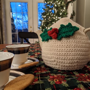May include: A white crocheted teapot cozy with green holly leaves and red berries. The cozy is on a green and red plaid tablecloth with a Christmas tree in the background.